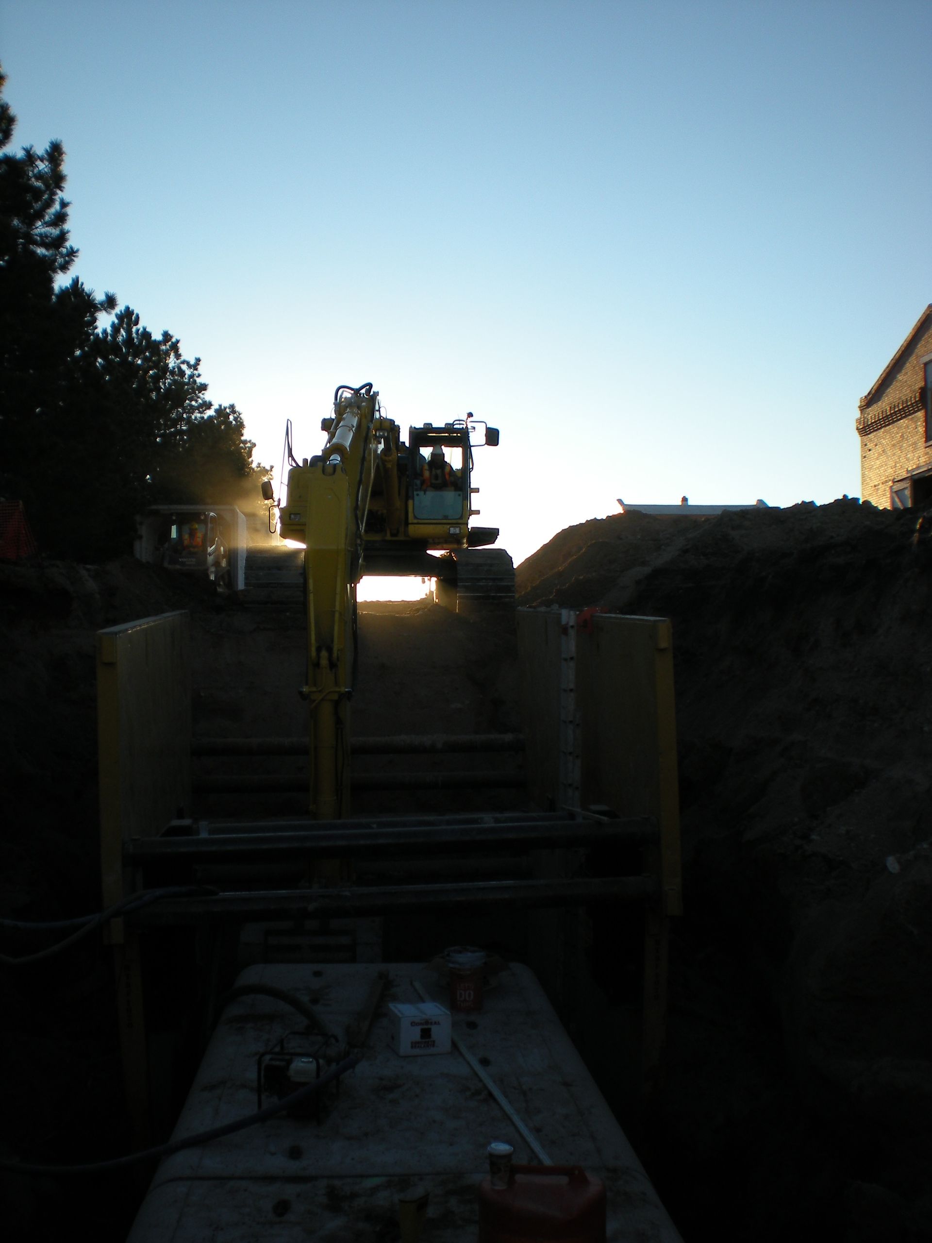 Construction machinery at dusk on a narrow site, with a silhouetted worker and trees framing the scene.