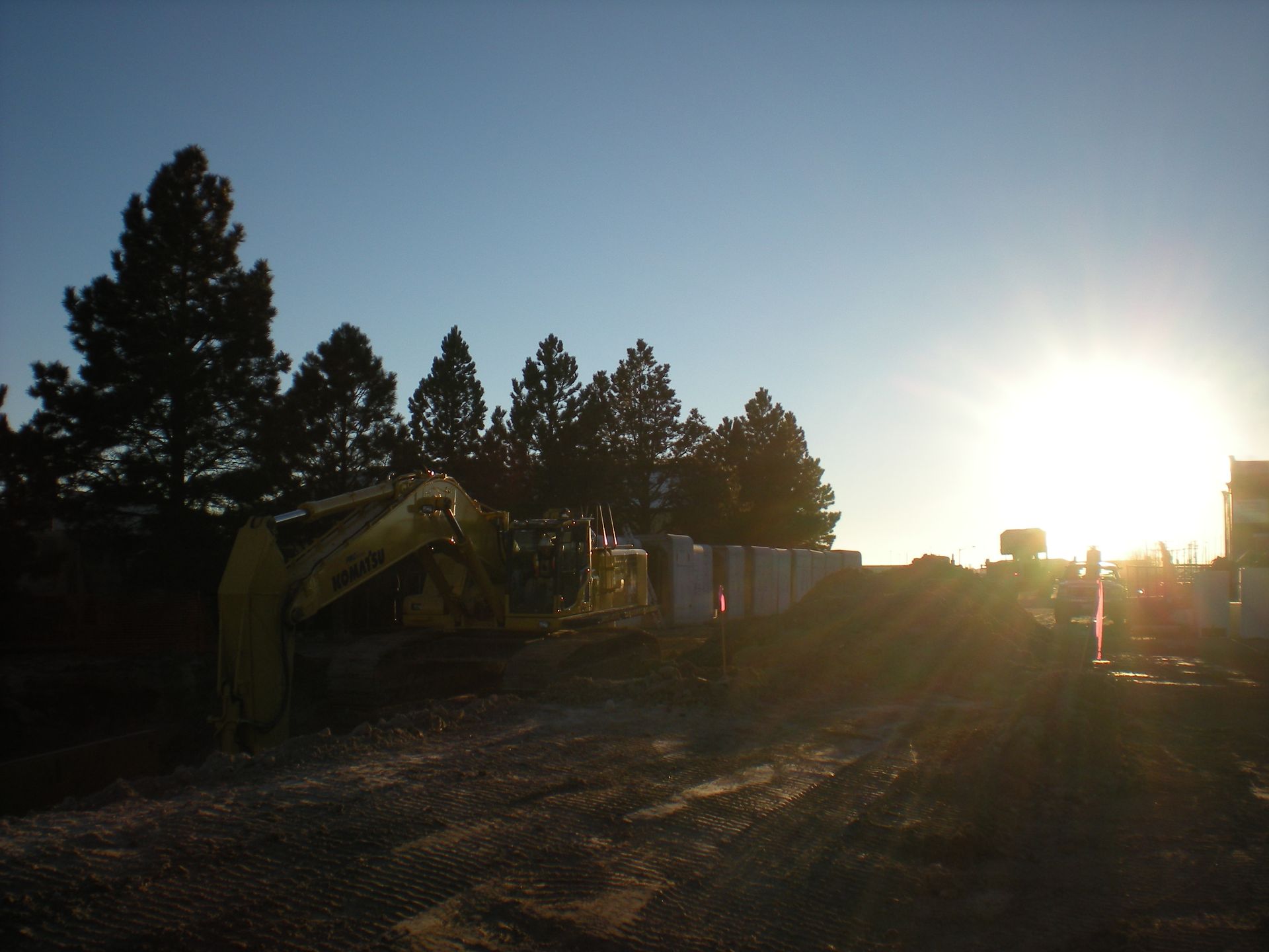 Sunlit construction site with excavator, dirt road, and tall trees silhouetted against the sky