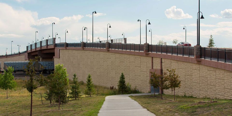 Elevated bridge with tan retaining wall, streetlights, and trees beside a grassy embankment under a blue sky