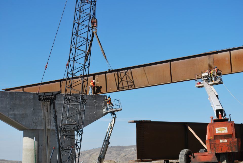 Construction workers on cranes installing a large steel bridge beam under a blue sky