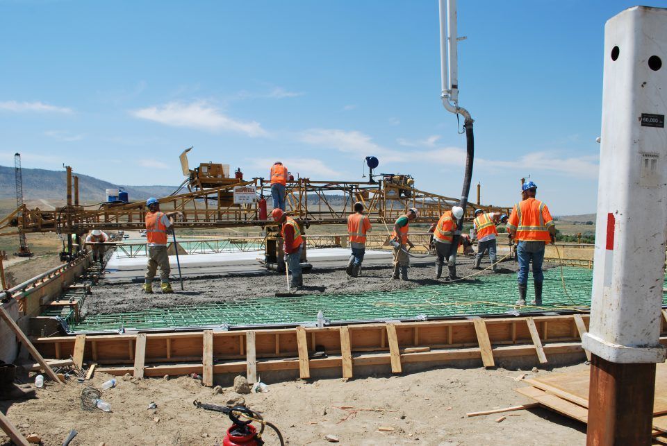 Construction workers in orange vests pouring concrete at a site with wooden forms and machinery