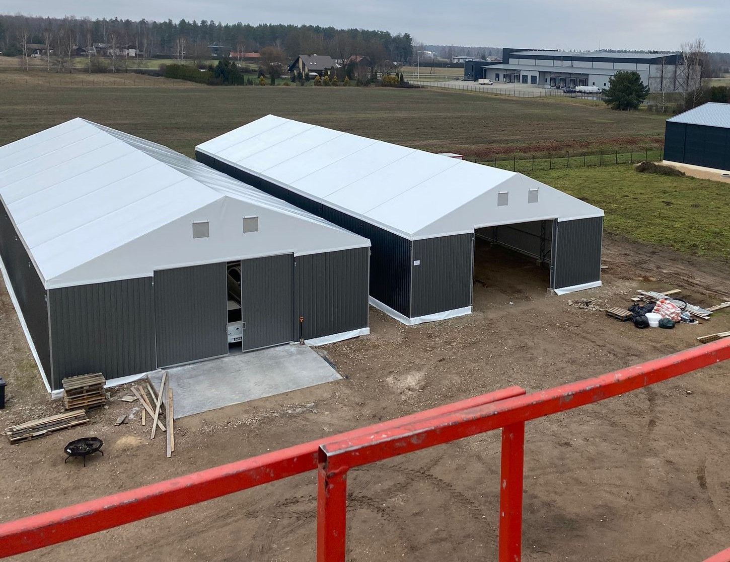 Du tamsiai pilki metaliniai sandėliavimo pastatai su baltais stogais kaimo aplinkoje, vaizdas iš viršaus.
