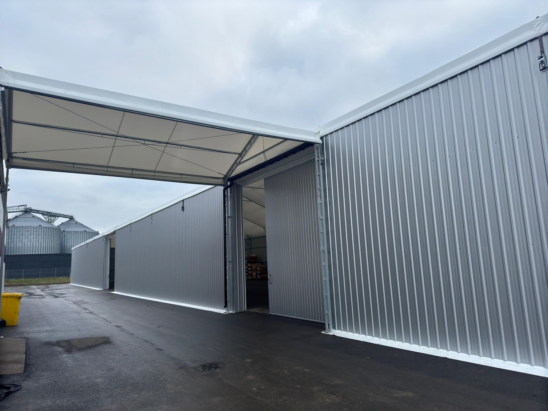 Ilgas, sidabrinis gofruoto metalo sandėlis su atviru įėjimu po baltu baldakimu, pilkas dangus.