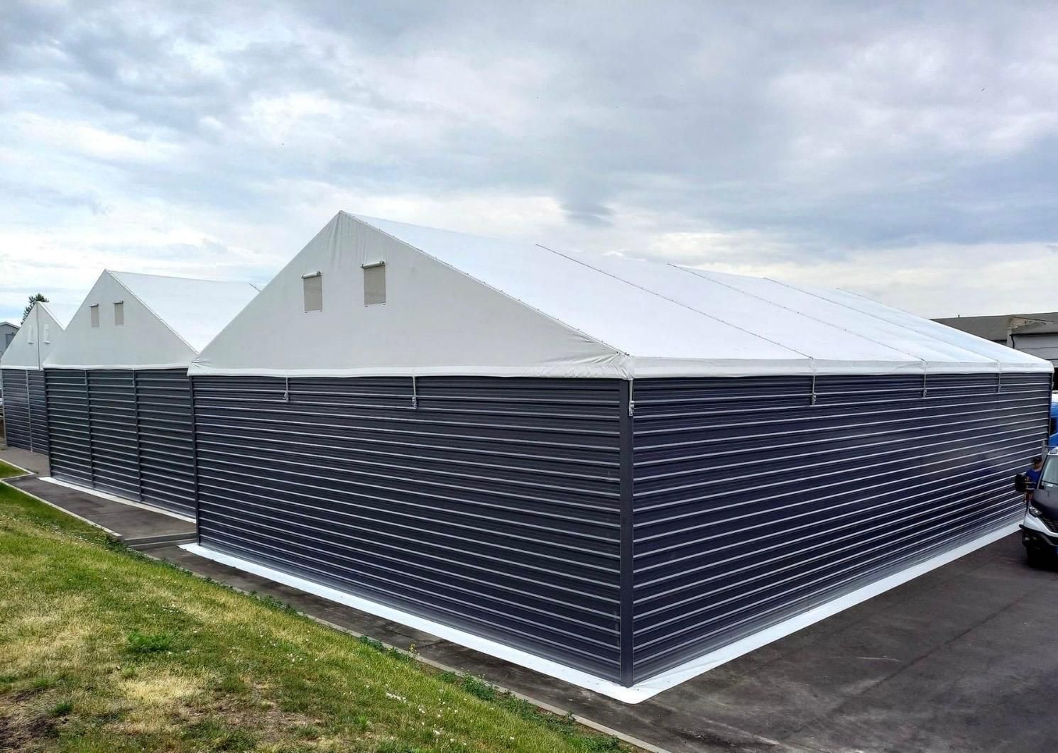 Trys dideli, tamsiai pilki gofruoto metalo pastatai su baltais šlaitiniais stogais po debesuotu dangumi.