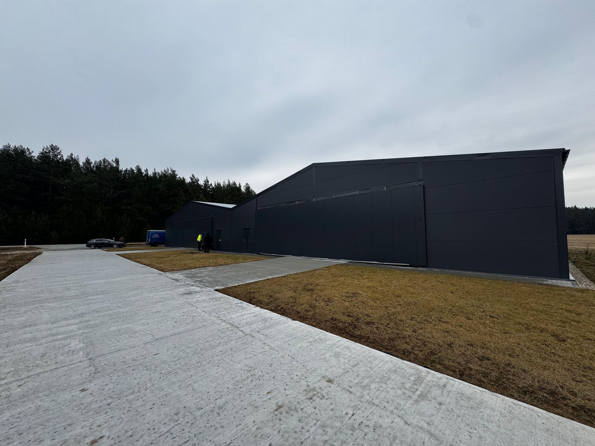 Pilkas pramoninis pastatas su betoniniu įvažiavimu, lauku ir medžių linija po debesuotu dangumi.