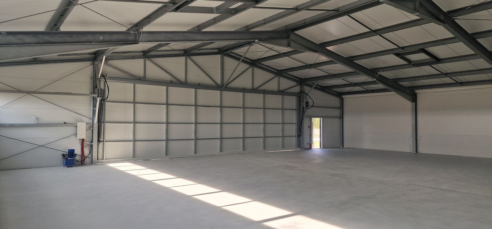 Tuščias pramoninis sandėlis, betoninės grindys, metalinis rėmas ir sienos, durys tolimajame gale.