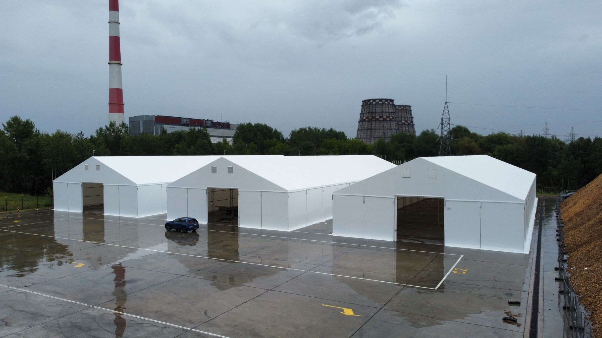 Trys balti laikini statiniai su atviromis durimis ant šlapio betoninio paviršiaus su tolimu pastatu ir kaminu.