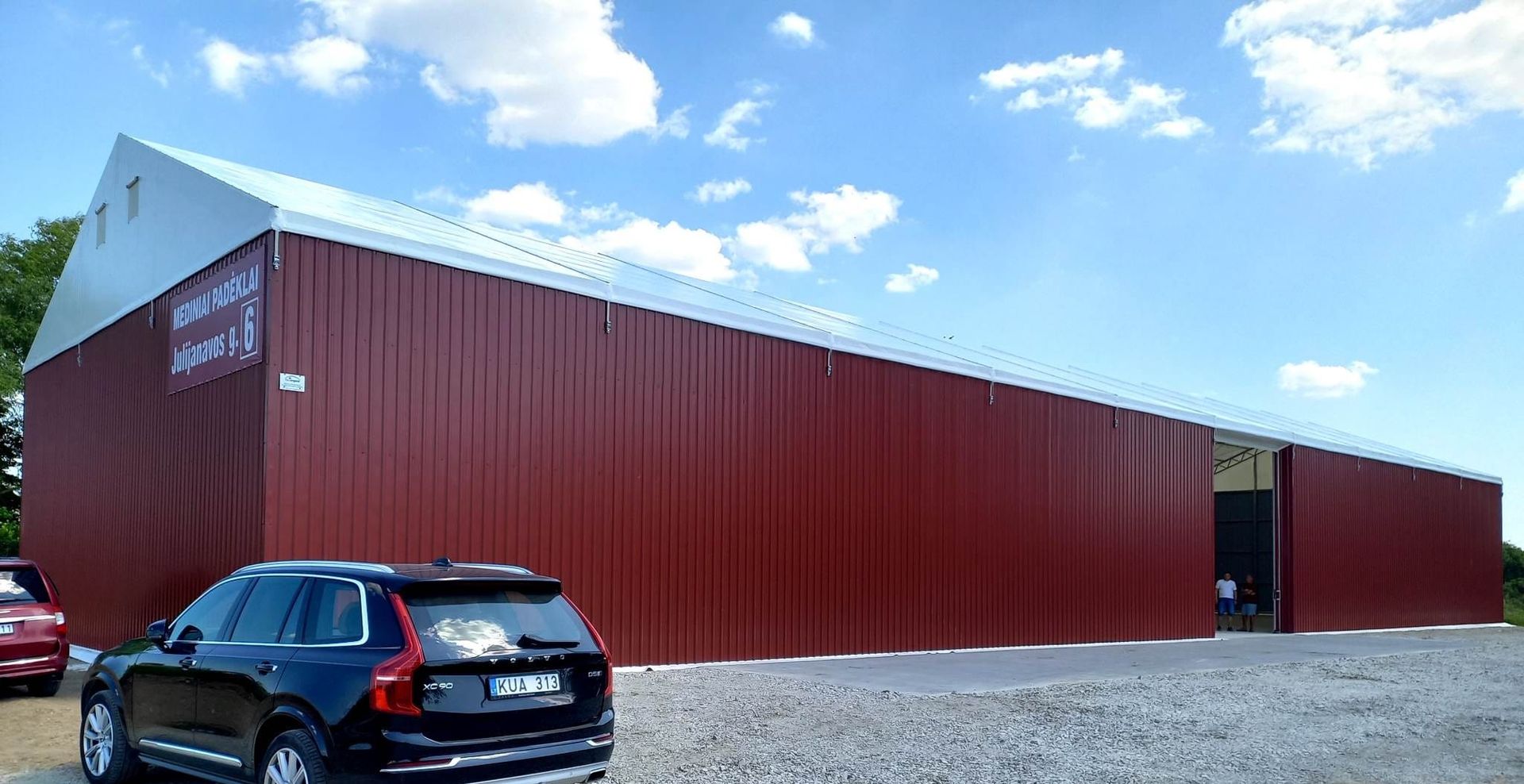 Raudonas tvartą primenantis pastatas su sidabriniu stogu po mėlynu dangumi. Priekyje pastatytas juodas visureigis.