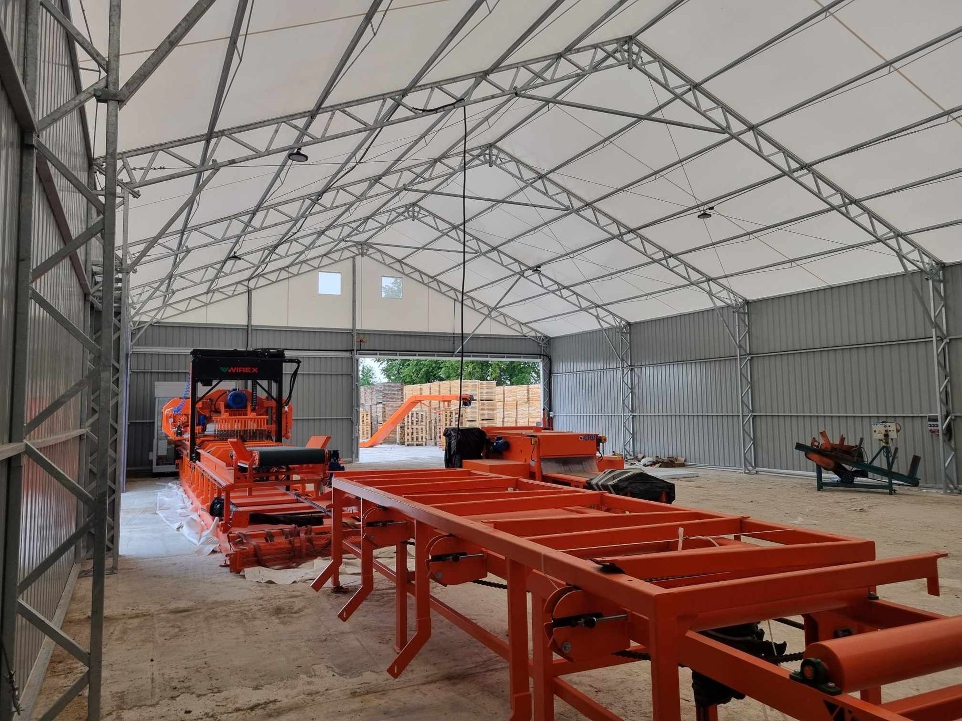 Oranžinės lentpjūvės įrenginiai didelėje metalinėje ir baltoje palapinę primenančioje konstrukcijoje.