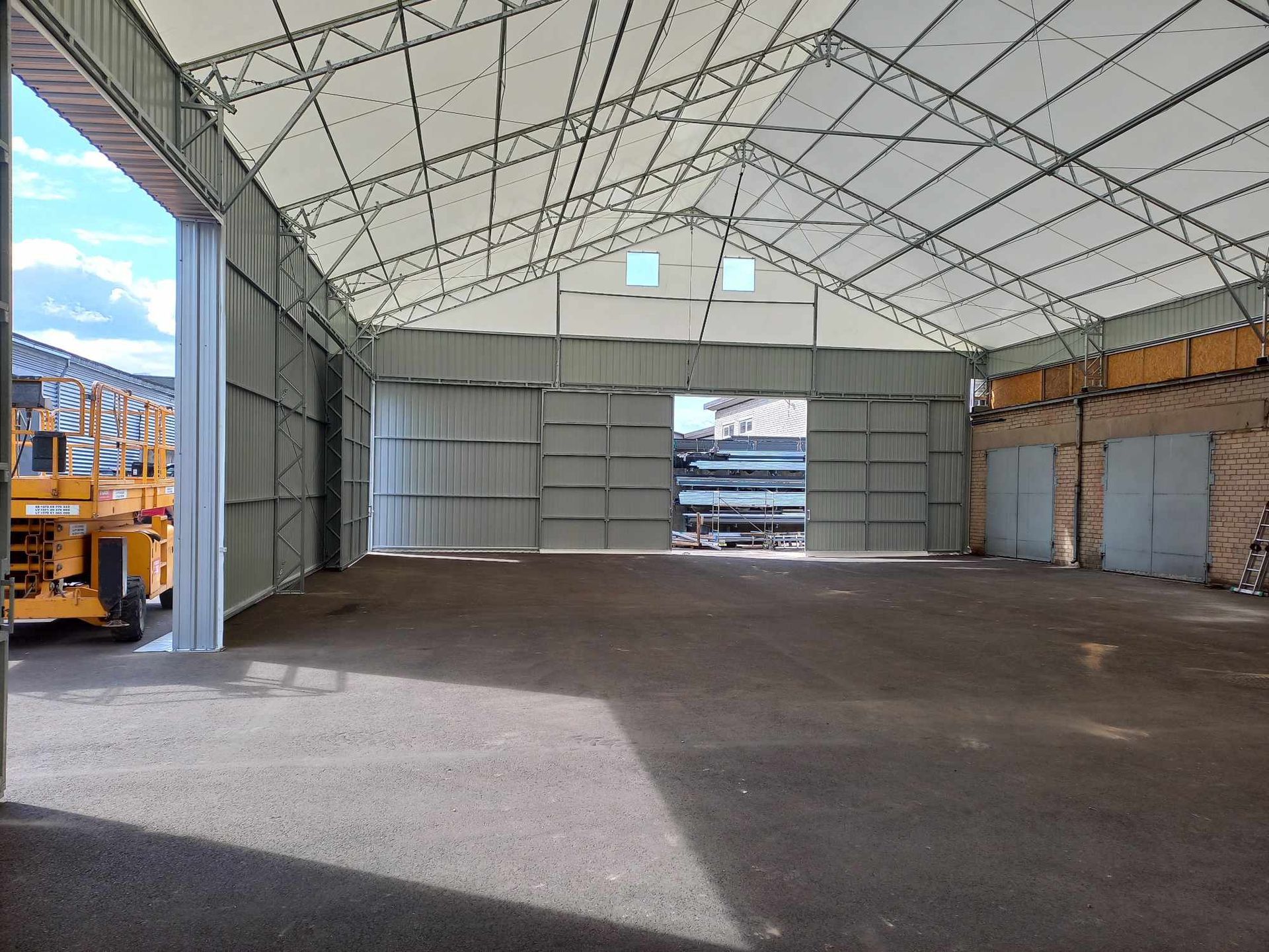 Didelio, atviro metalinio sandėlio vidaus vaizdas. Saulės šviesa sklinda pro dideles atviras duris.