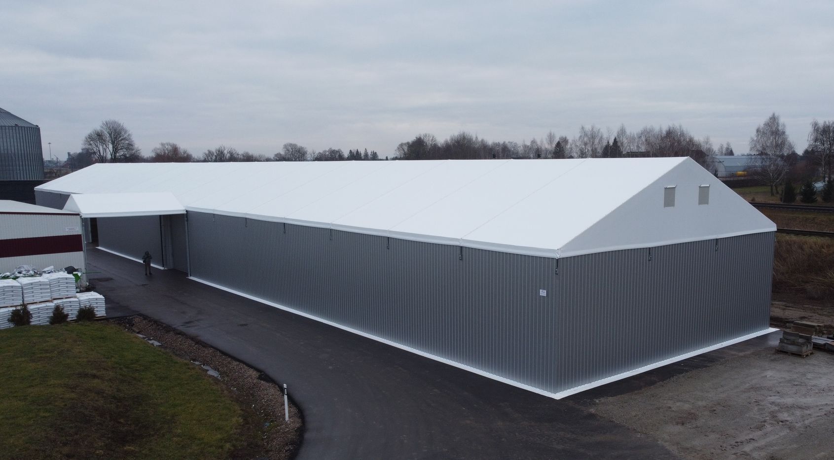 Sandėlis su baltu stogu ir pilkais gofruotais šonais debesuotą dieną.