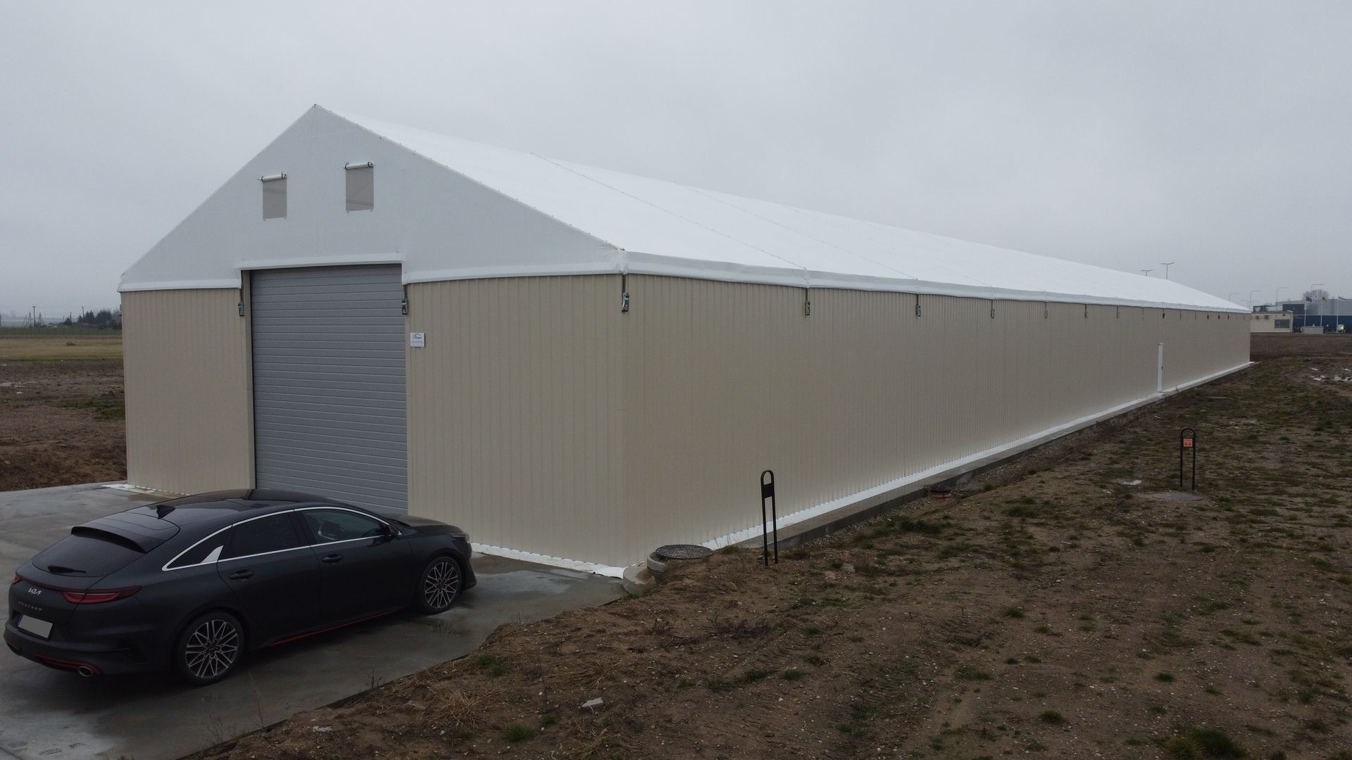 Smėlio ir baltos spalvos pramoninis pastatas su priekyje pastatytu automobiliu. Apsiniaukusi diena.