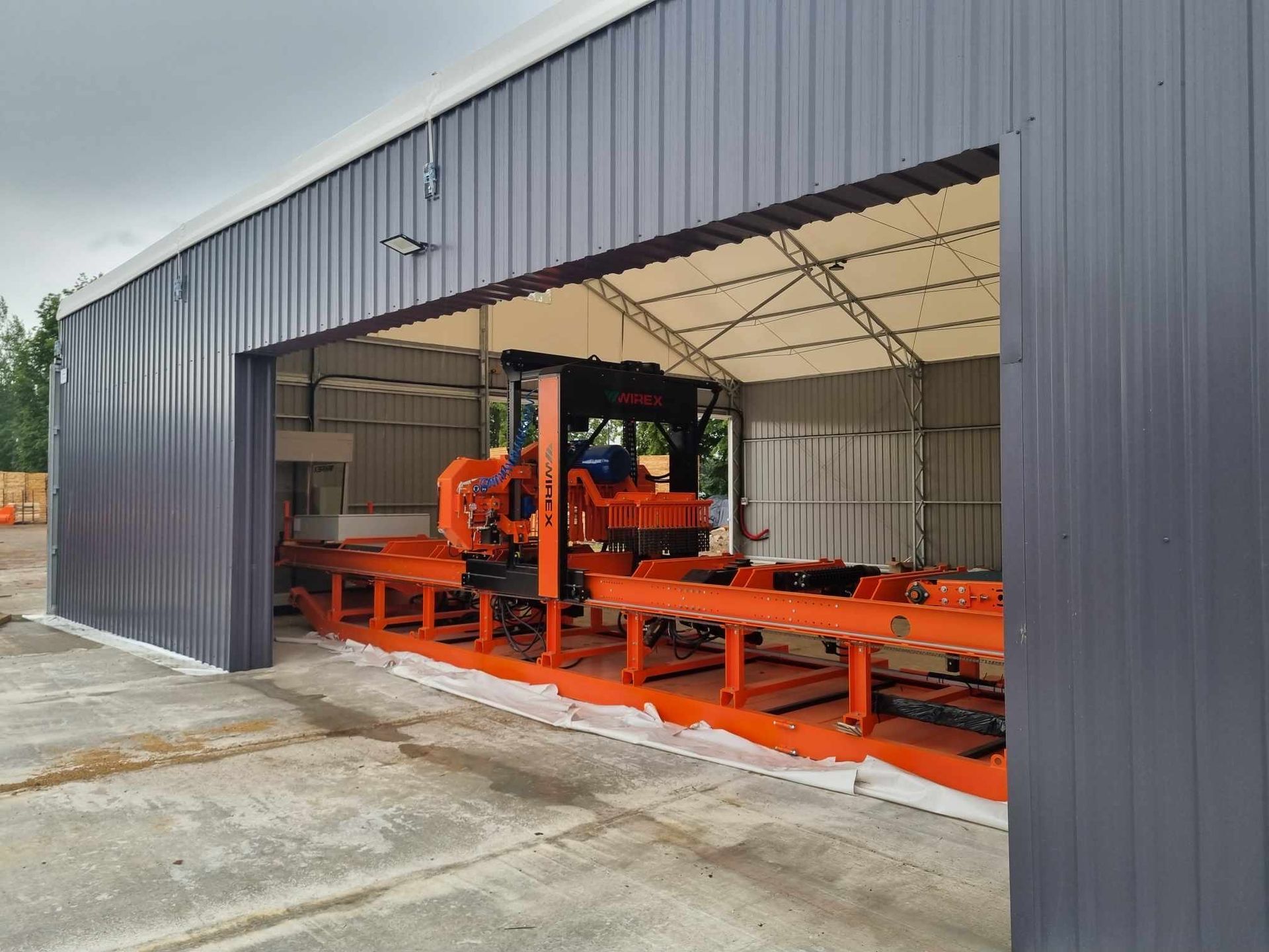 Lentpjūvė pilkame metaliniame angare. Mechanika oranžinė. Fotografavimo vieta – lauke.