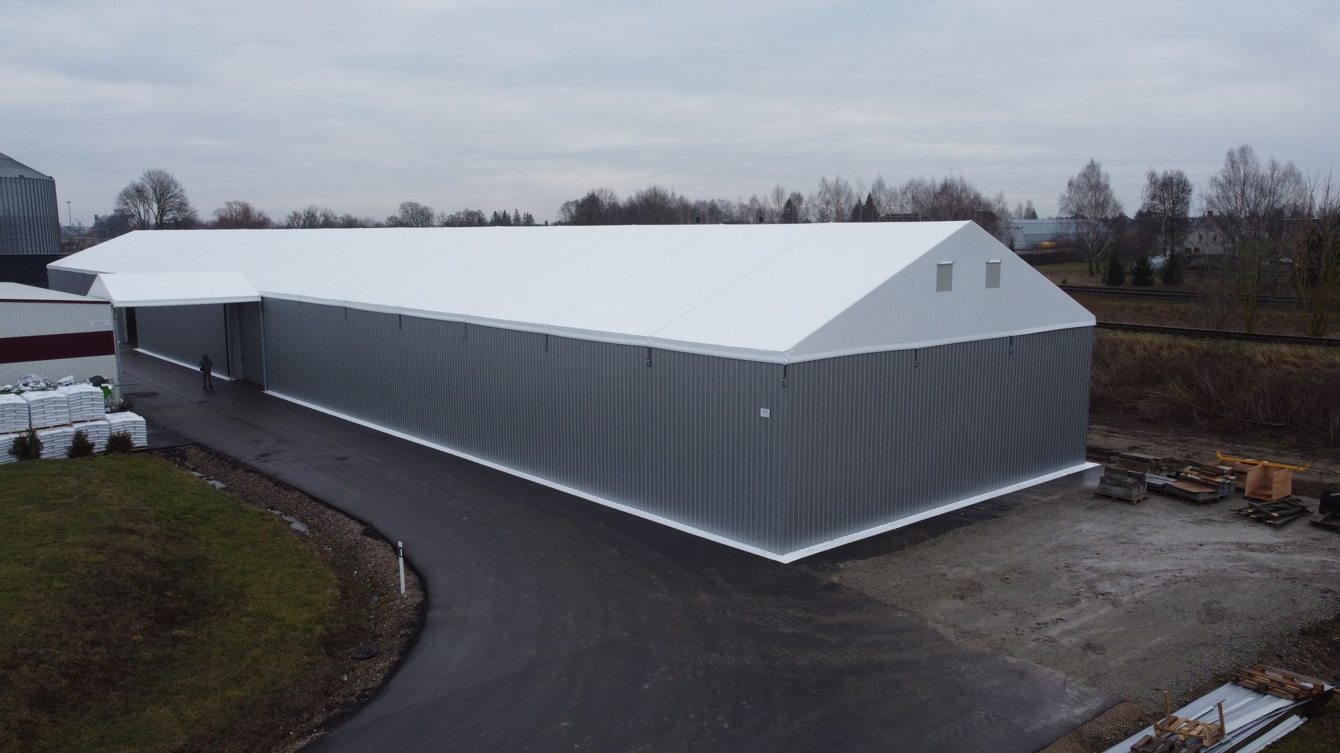 Pilkas pramoninis sandėlis su baltu stogu debesuotą dieną.
