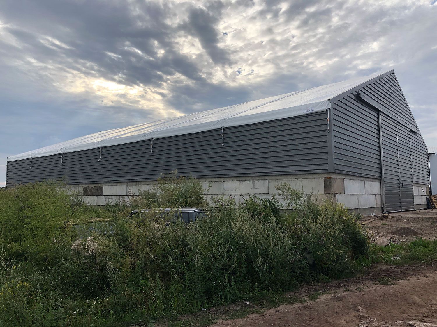 Didelis pilkas pramoninis pastatas su kampuotu stogu debesuoto dangaus fone, apsuptas apaugusios žalumos.