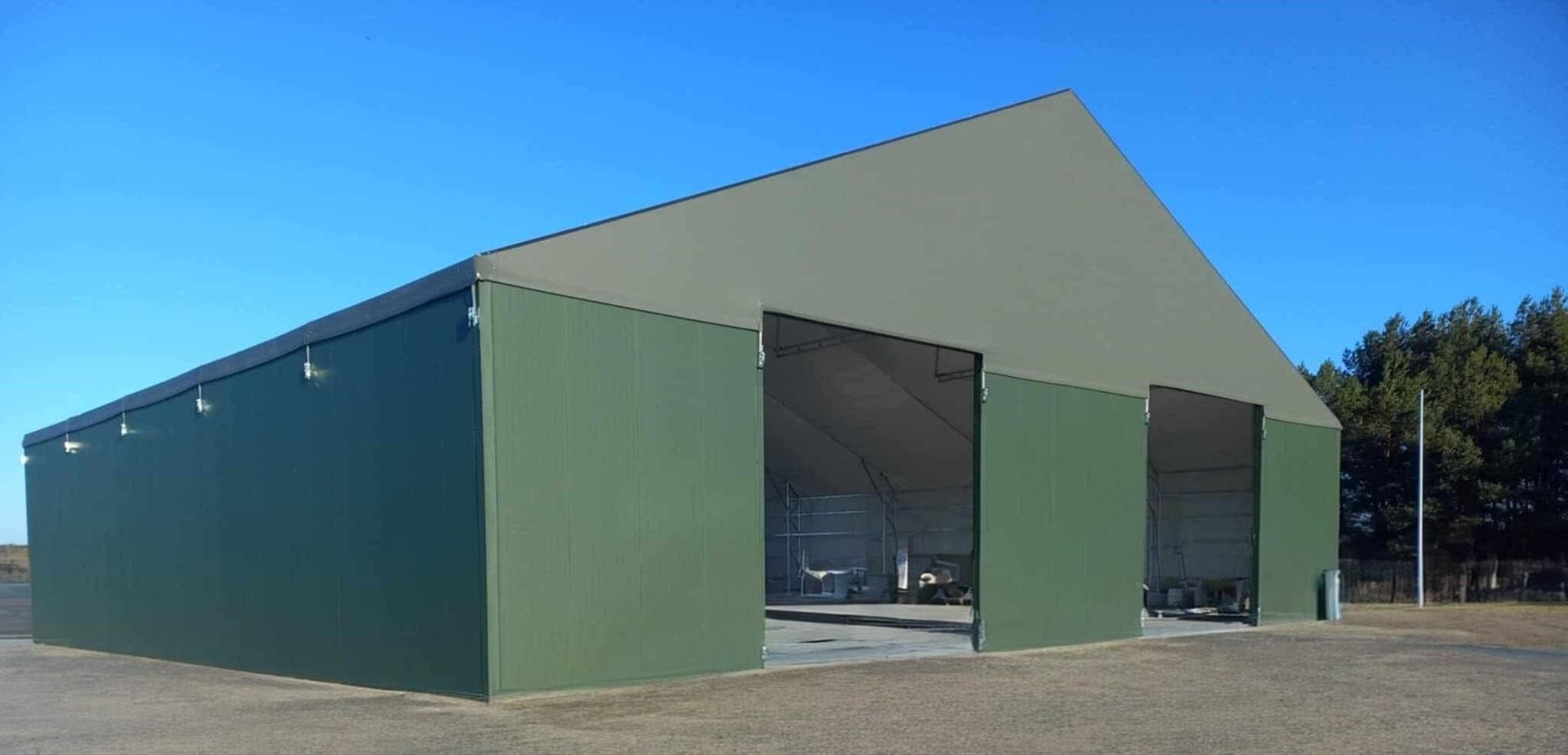 Žalias ir gelsvai rudas metalinis žemės ūkio pastatas su didelėmis atviromis durimis giedro mėlyno dangaus fone.