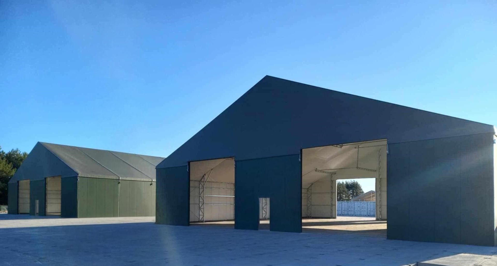 Du dideli žali sandėliai su atviromis durimis saulėtą dieną.