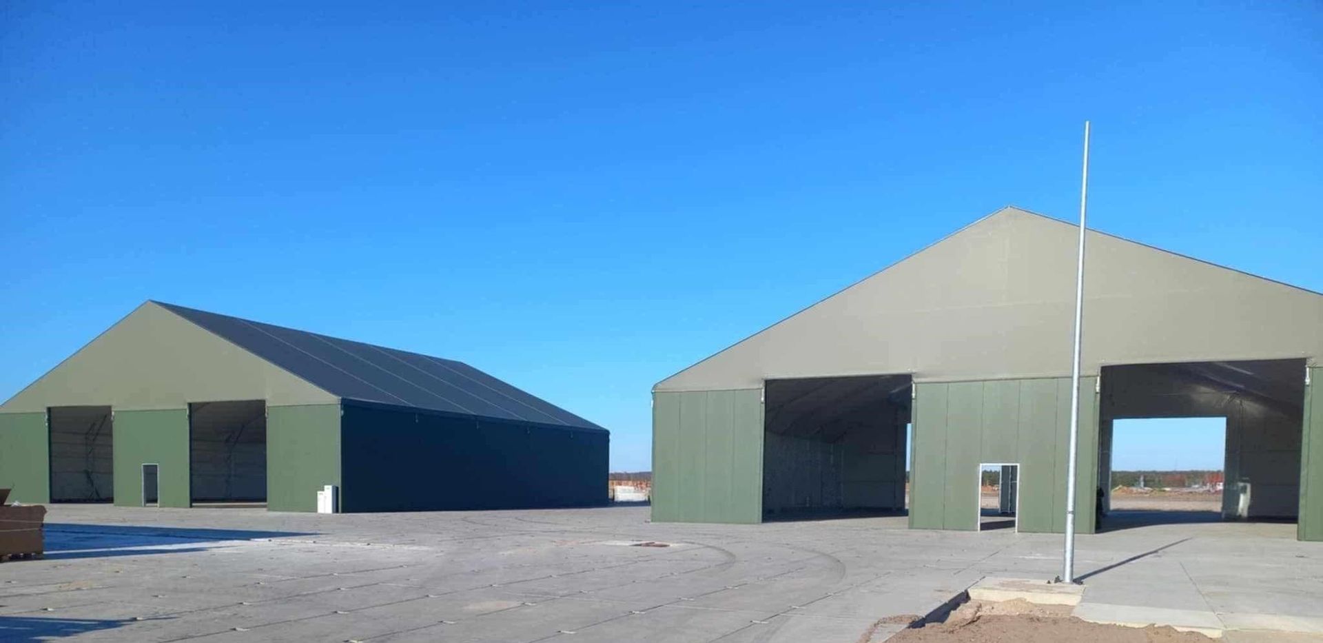 Du žali pastatai su atviromis durimis giedrą, mėlyną dieną. Priekyje stovi vėliavos stiebas.