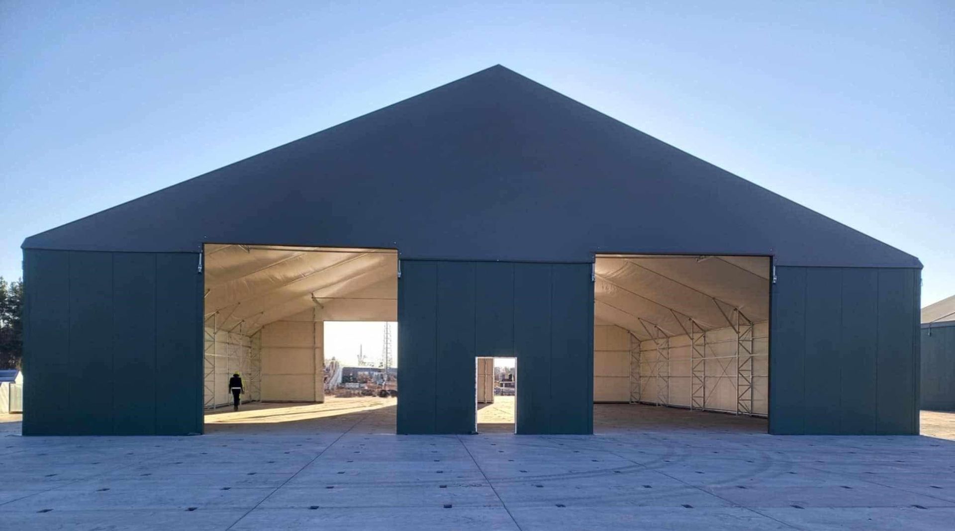 Didelis, pilkas, atvirų kraštų pastatas su dvišlaičiu stogu ant betoninio paviršiaus po giedru dangumi.