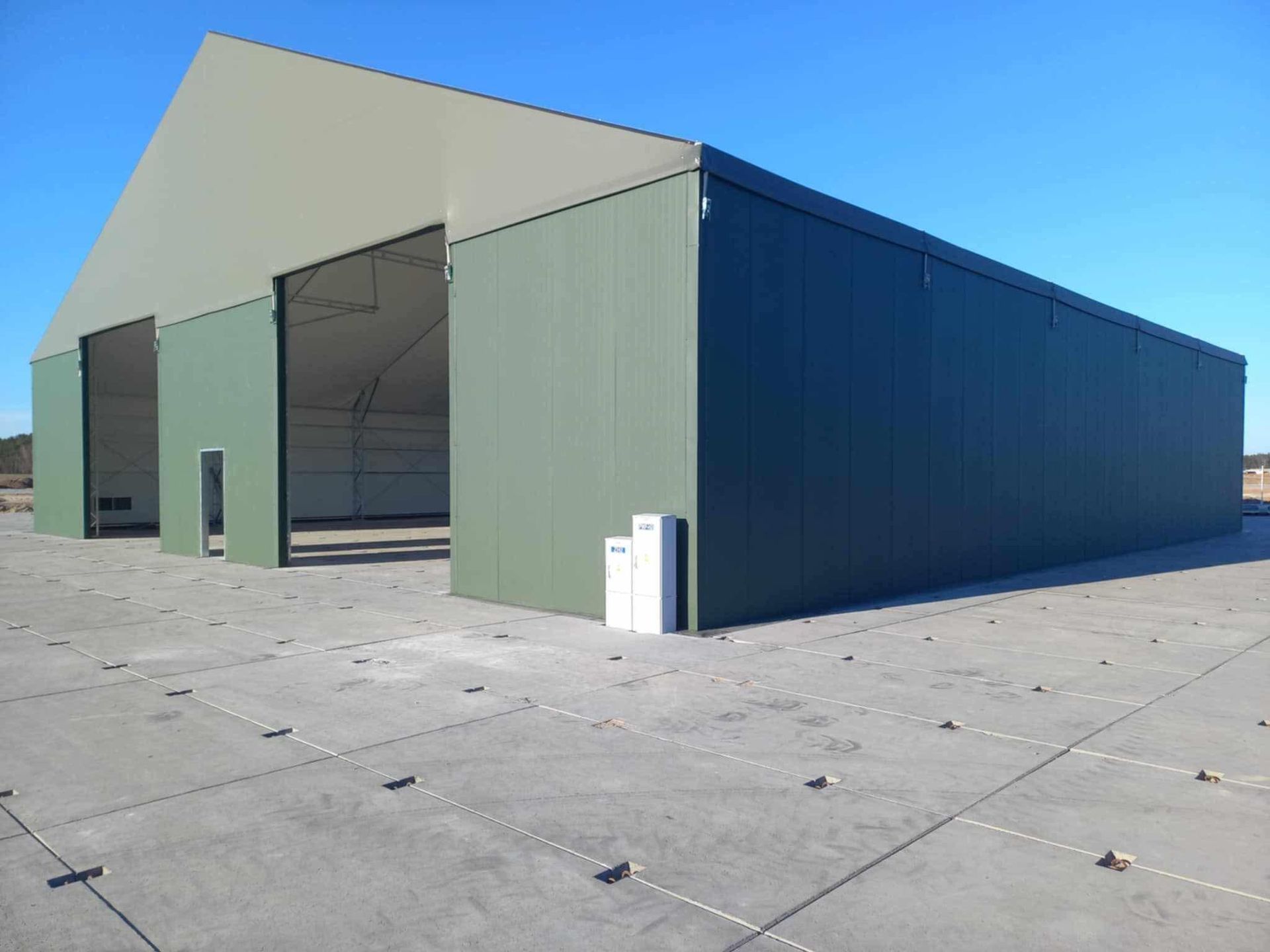 Didelis žalias metalinis angaras su atviromis durimis ant betono.