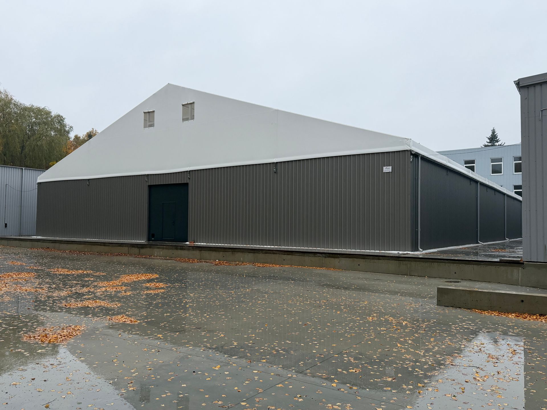 Pilkas metalinis sandėlis su baltu kampuotu stogu. Durys ir maži langeliai stoge. Išorinė aplinka, apsiniaukęs dangus.