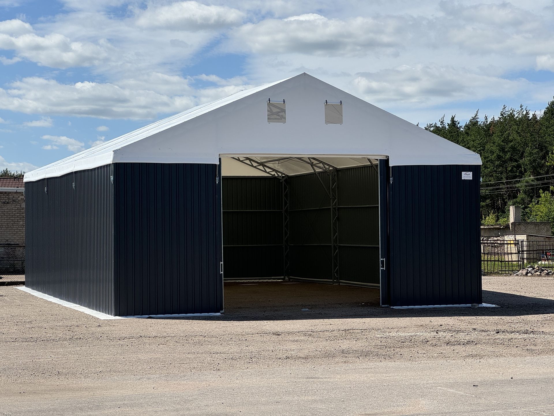 Tamsiai pilkas metalinis sandėliukas su baltu stogu ir atviromis durimis ant žvyro.