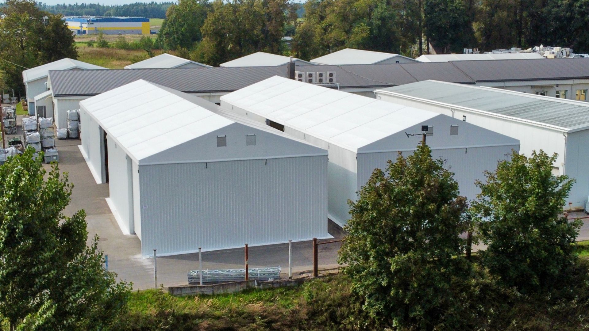 Baltos sandėlio palapinės objekte, apsuptame medžių ir pastatų po mėlynu dangumi.