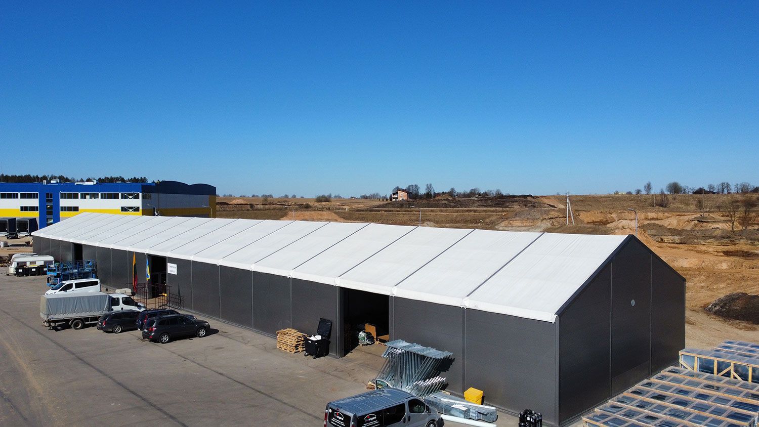 Sandėlis su transporto priemonėmis ir žmonėmis lauke po giedru mėlynu dangumi.