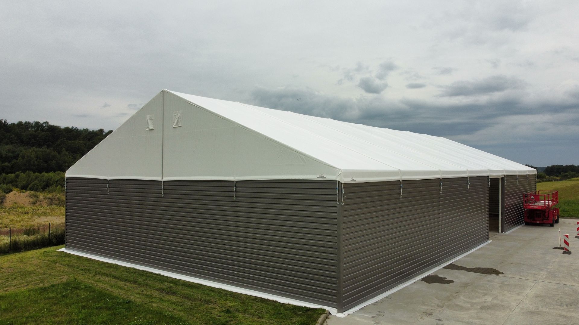 Pilkas ir baltas pramoninės palapinės tipo pastatas su šlaitiniu stogu, pastatytas žolėtame lauke po debesuotu dangumi.