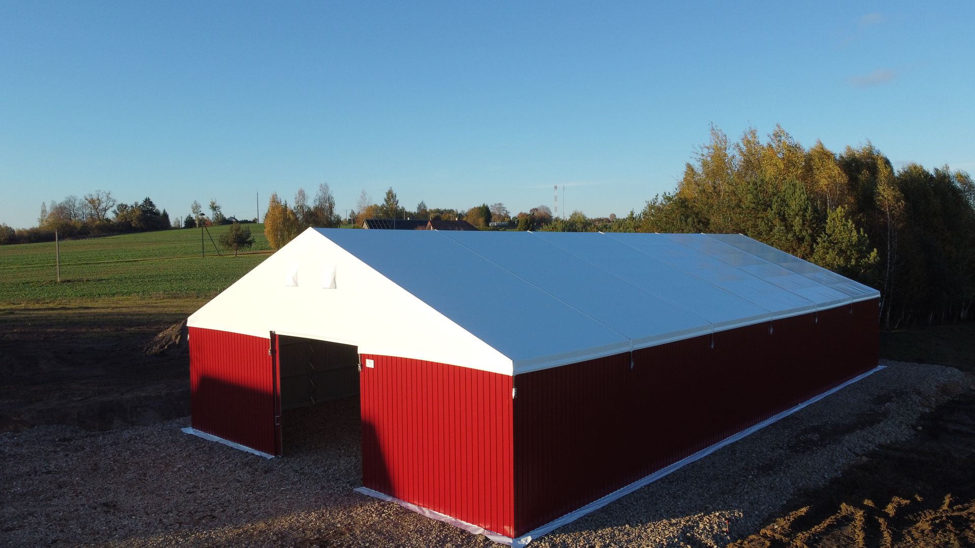 Raudonai baltas metalinis tvartas kaimo aplinkoje, žvyruotas žemės paviršius, mėlynas dangus, medžiai fone.