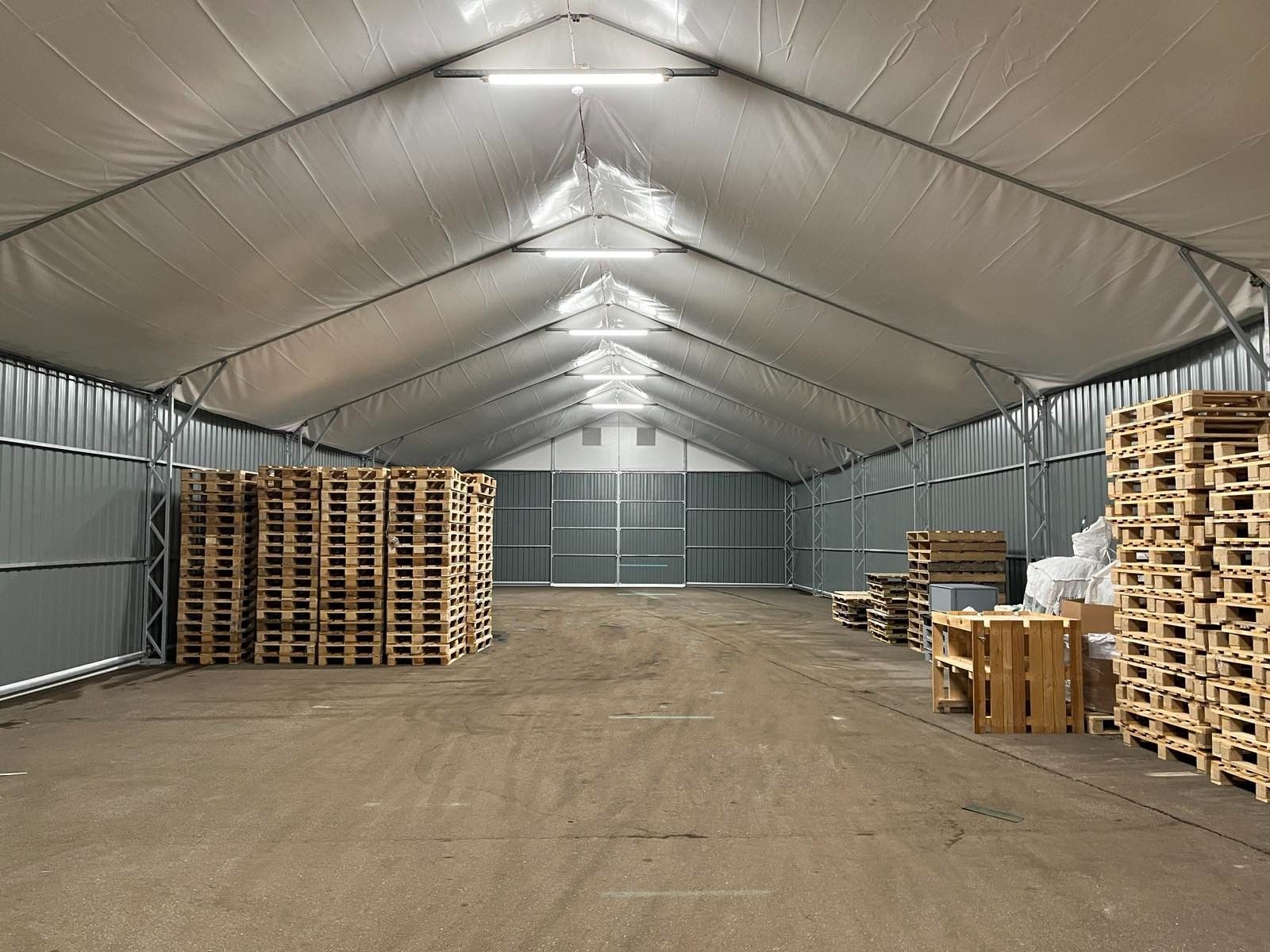 Didelio sandėlio su medinių padėklų krūvomis ir metalinėmis dailylentėmis vidaus vaizdas; ryškūs viršutiniai žibintai.