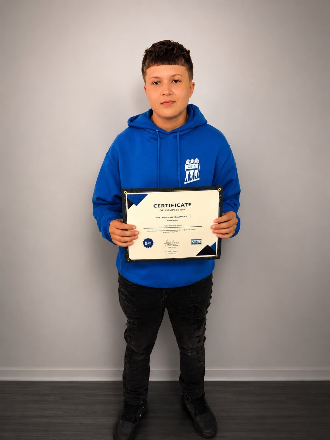 Person in a blue hoodie holding a certificate against a plain gray wall.