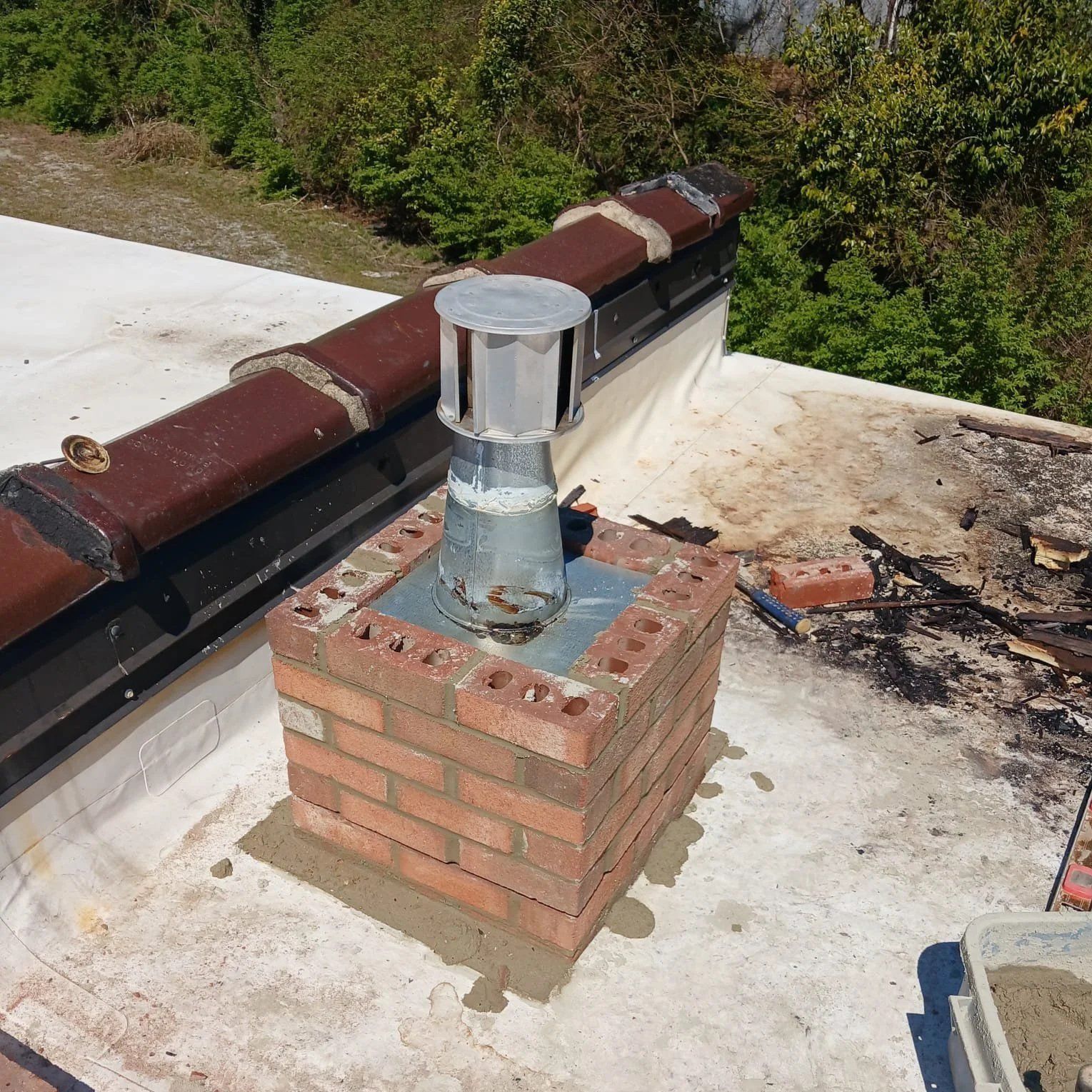 Brick chimney vent on a flat roof with rusted metal flashing and surrounding debris.