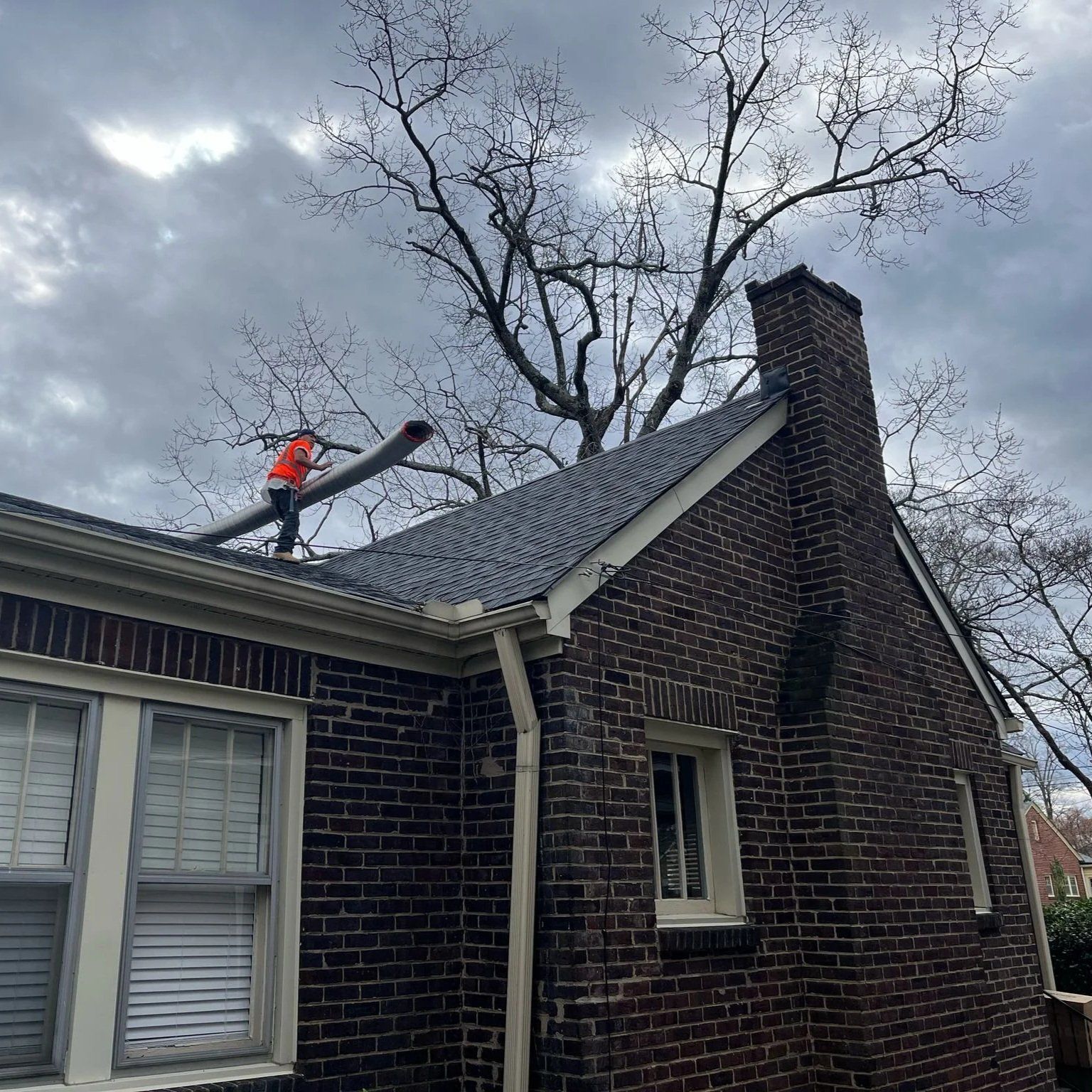 Worker in orange on a roof, near a brick chimney under a cloudy sky.