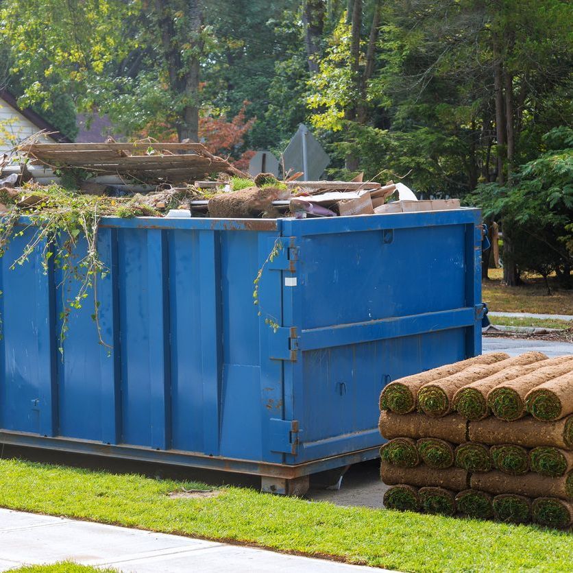 A blue dumpster is sitting on the side of the road next to a pile of turf.