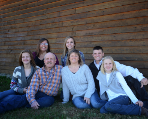 Seven people sitting on grass in front of a wooden wall, smiling at the camera.