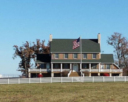 A farmhouse with an American flag, white fence, and leafless trees on a sunny day.