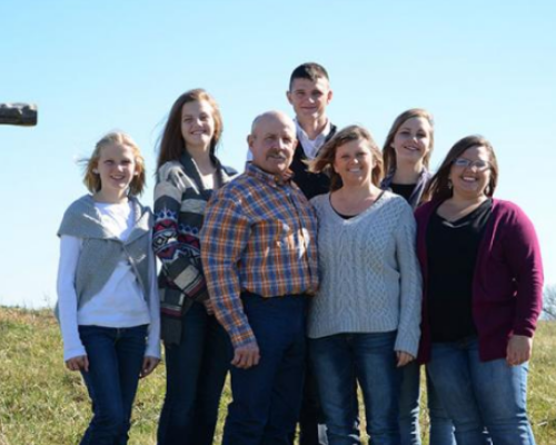 Six people standing together outdoors in a sunny field, smiling at the camera.