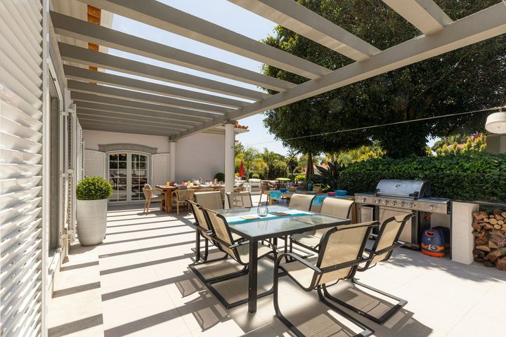 Covered patio with dining table, lounge chairs, and grill beside a house, lit by sunlight.