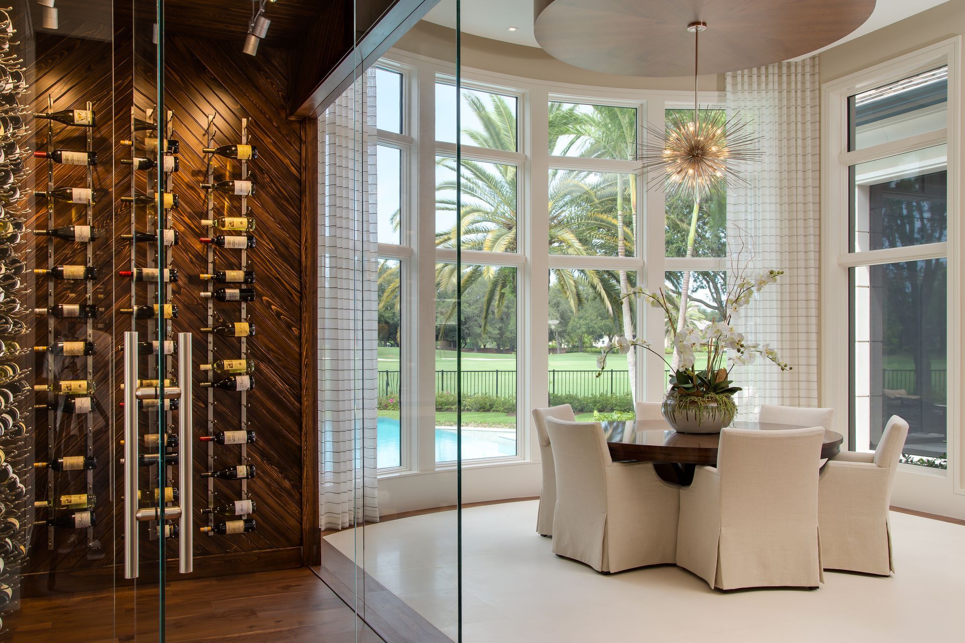 Dining room with wine storage, large windows overlooking a pool and palm trees. Neutral tones, elegant.