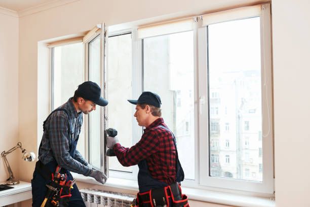 Two workers in caps and coveralls installing a window. One uses a drill. Bright indoor setting.