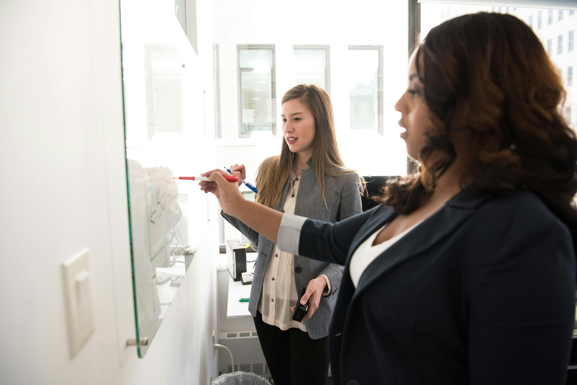 Two women in business attire at a whiteboard, one writing with a red marker, the other watching.