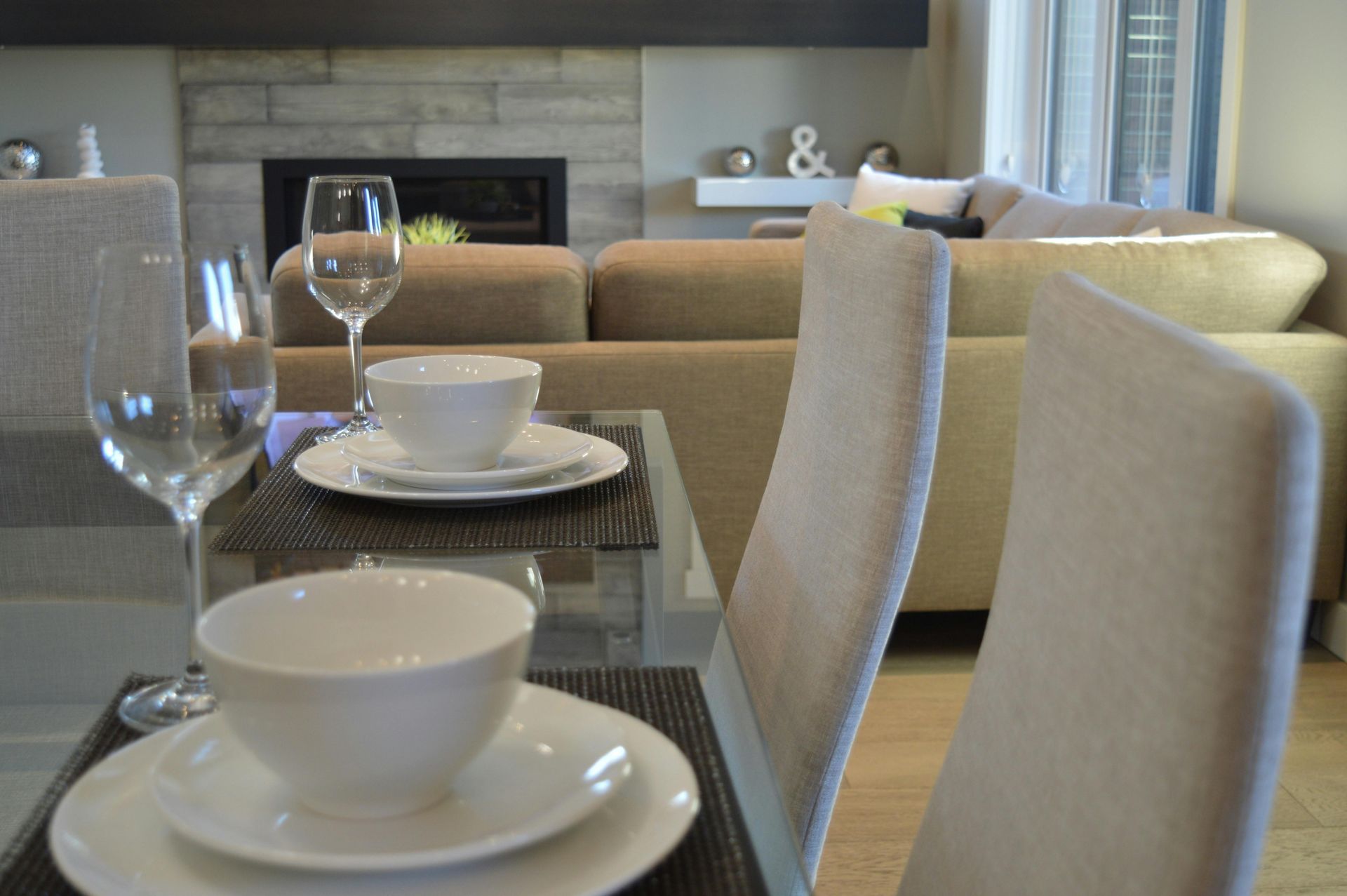 Glass Table. Dining table set for a meal, living room in the background. Glassware, white plates, beige chairs.