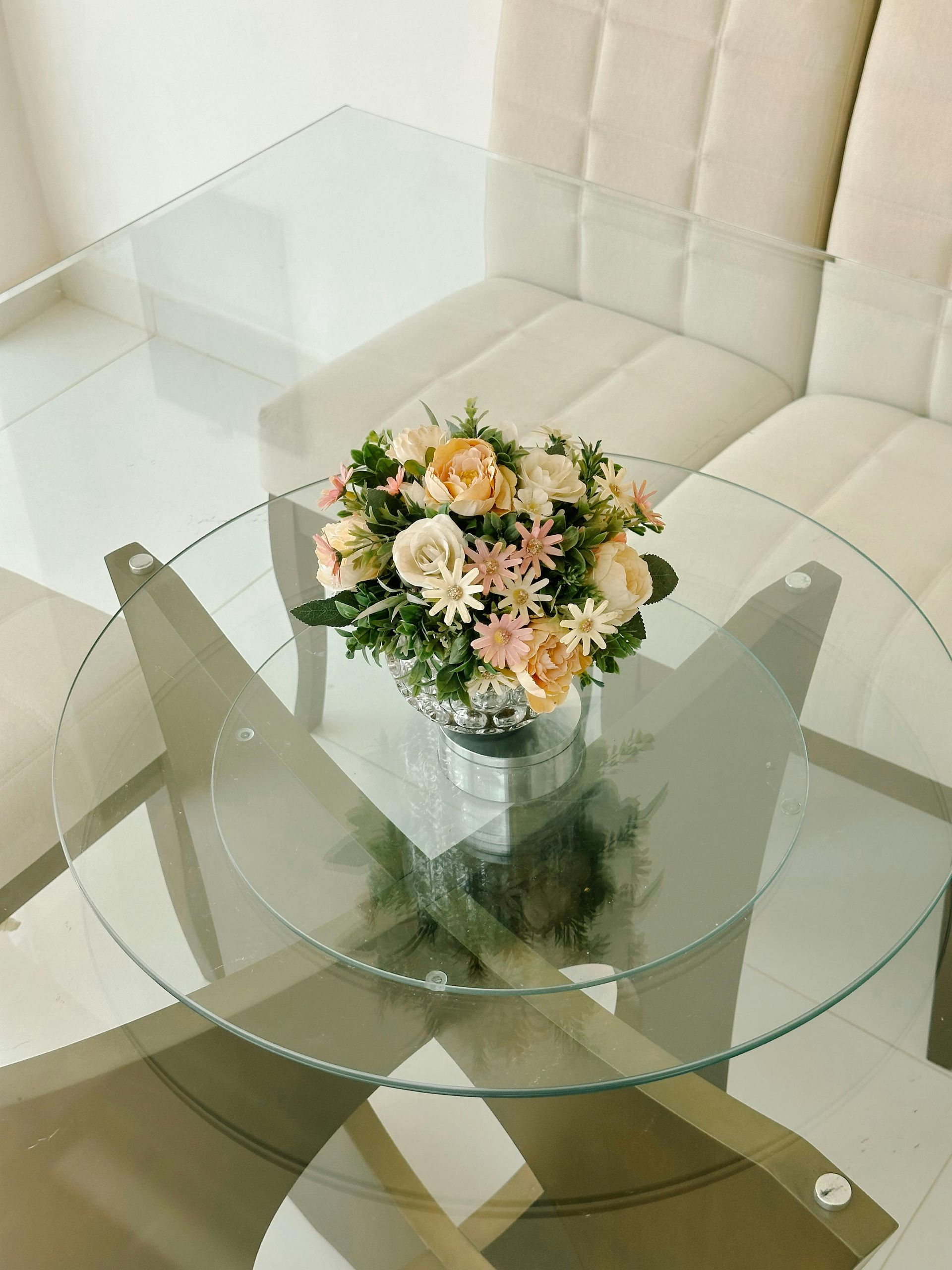 Glass dining table with floral centerpiece and ivory chairs.