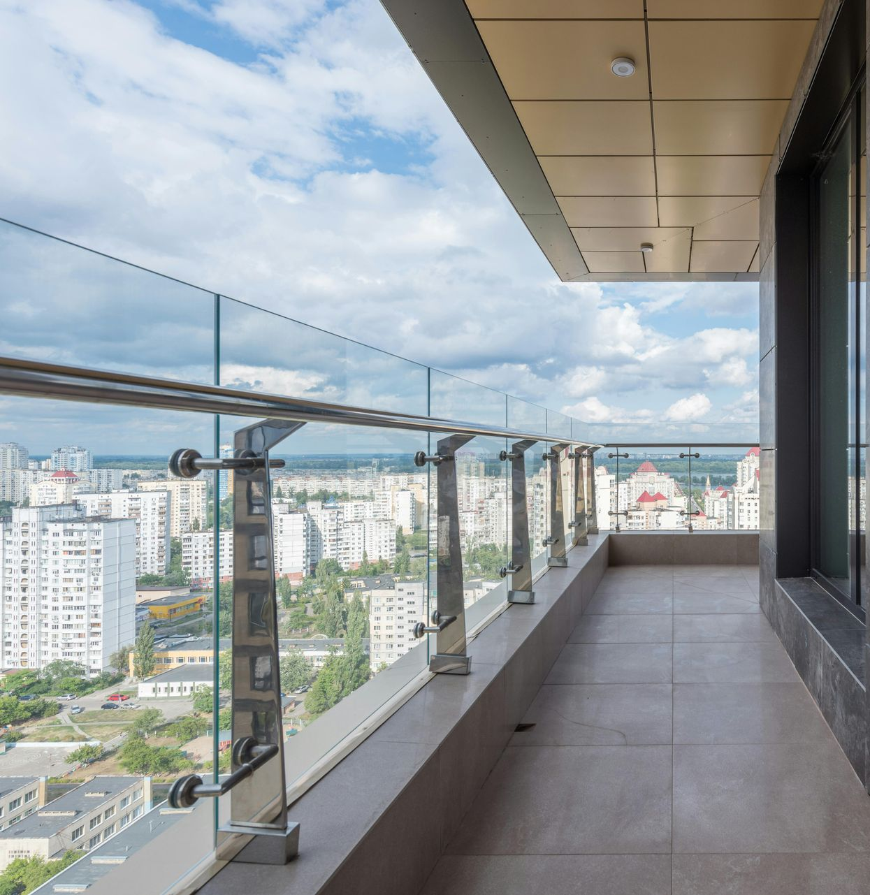 Balcony overlooking a cityscape, featuring glass railing, brown tile floor, and a cloudy blue sky.