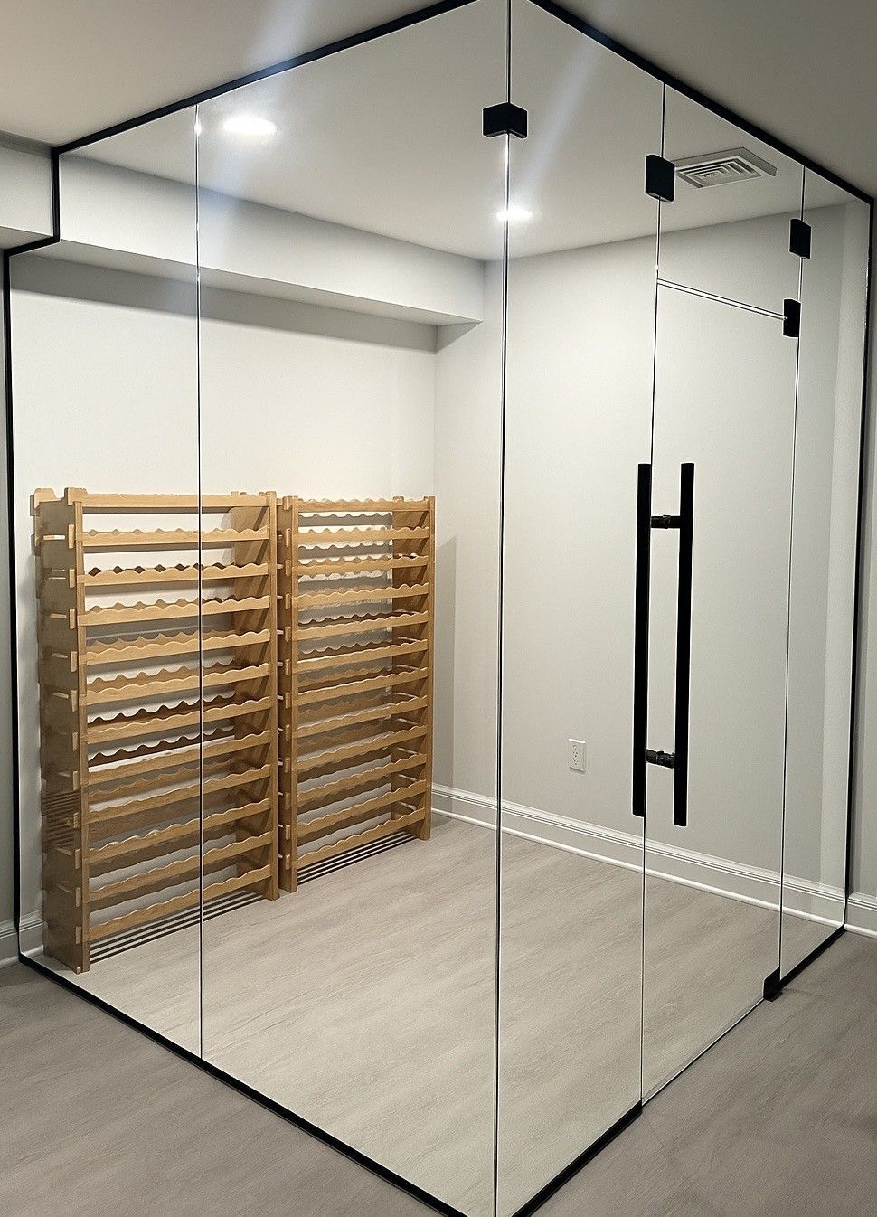 Glass-walled wine cellar with wooden racks holding bottles. Black door handle, gray flooring, and white walls.