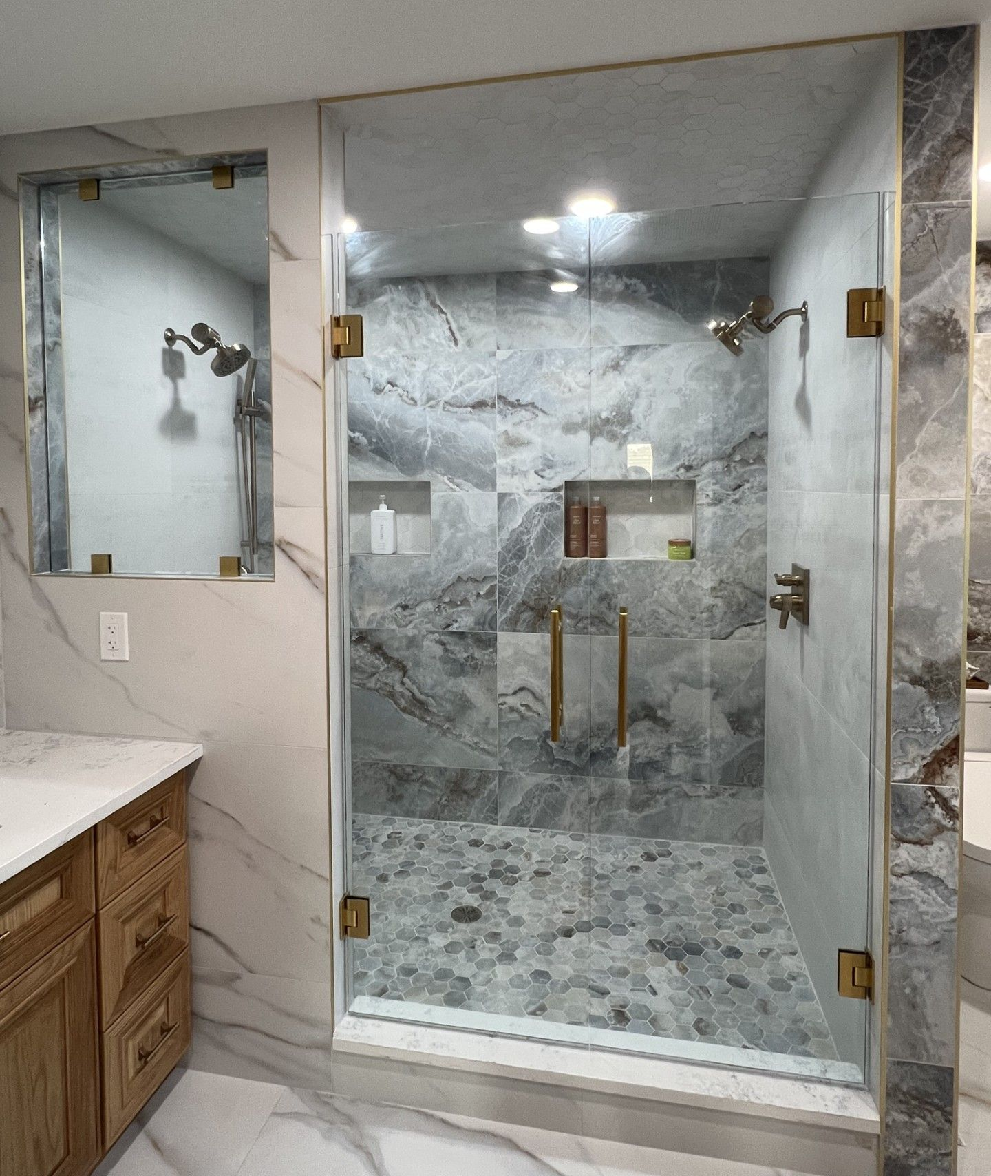 Glass shower enclosure with gray marble tile, golden hardware, and pebble floor. Oxford, MI