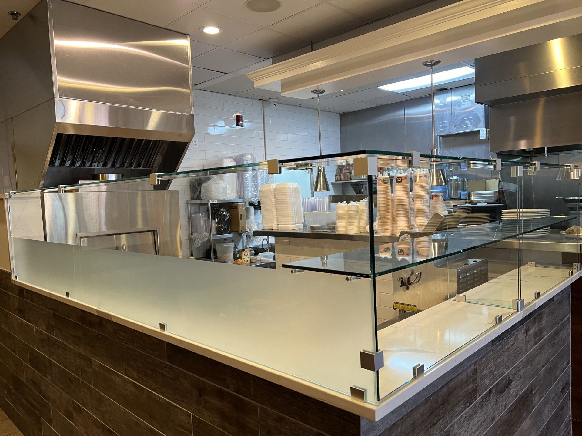Interior view of a commercial kitchen. Stainless steel equipment, glass counter, and brown wood paneling.