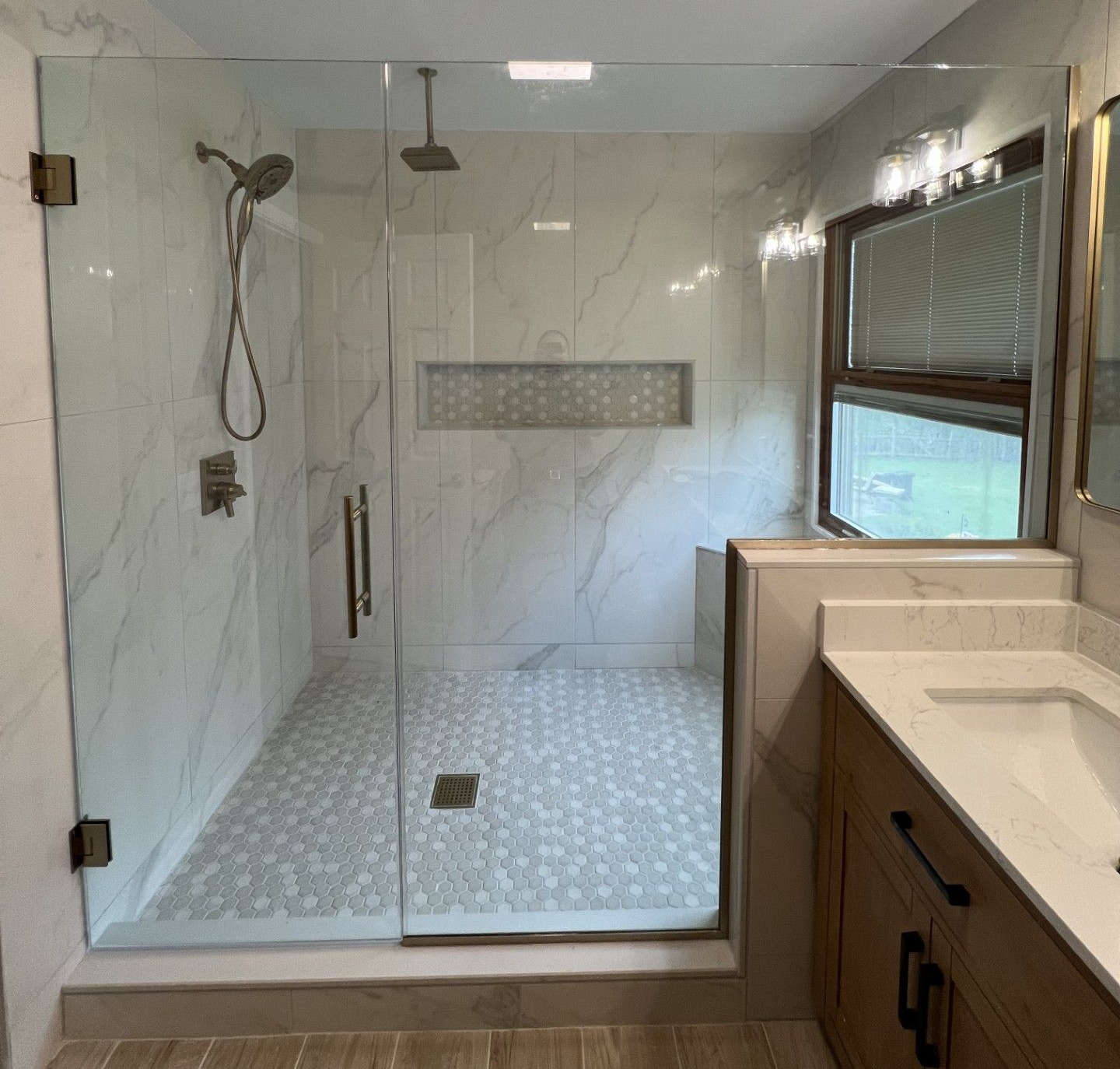 Bathroom with glass shower, white marble tile, pebble shower floor, wooden vanity, and window. Canton
