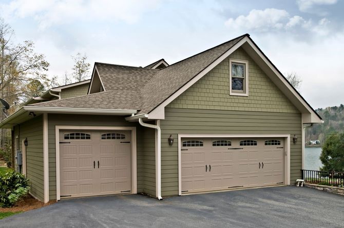 Two-car beige garage with tan doors and green siding on a sloped driveway near water